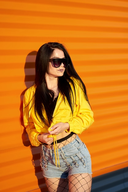 Moda jovem bonita vestindo juventude elegante casaco amarelo, shorts jeans e óculos de sol posando no fundo da parede urbana laranja.