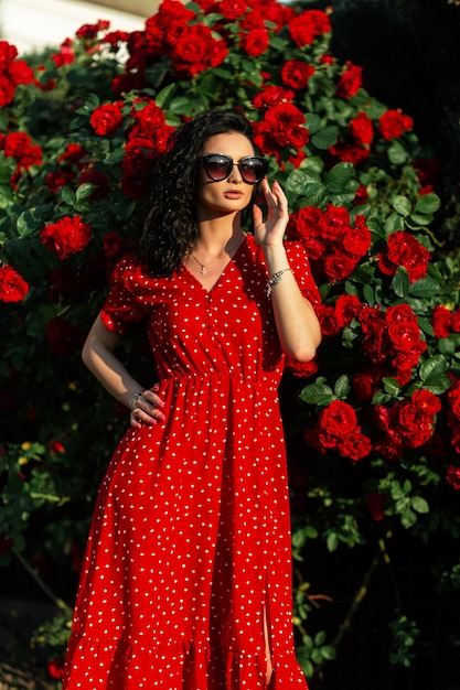 Moda jovem e linda mulher elegante com óculos de sol da moda em um vestido vermelho elegante se passando perto de um arbusto com flores de rosas