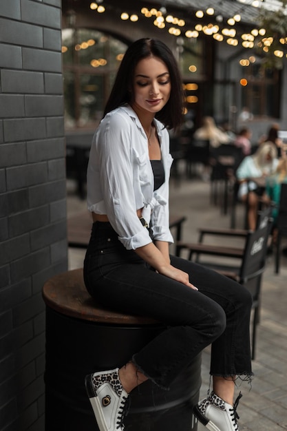 Moda jovem linda mulher com camisa branca e jeans preto com tênis senta-se no barril de metal preto na rua com luz bokeh