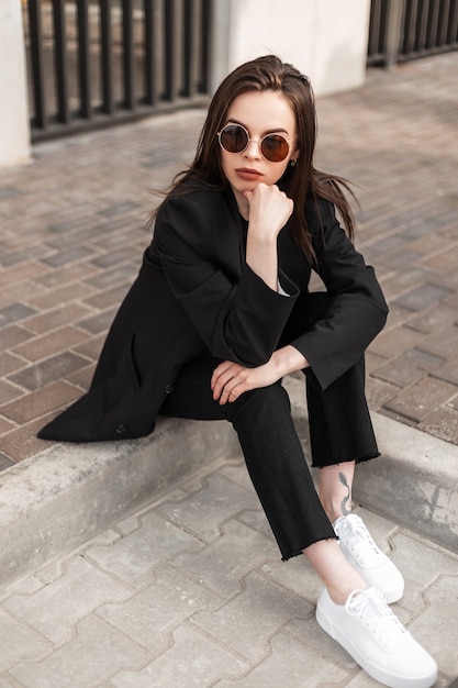 Moda jovem mulher em calças da moda em elegante blazer jovem sentado sobre ladrilhos de pedra na cidade. Nova coleção elegante de roupas femininas. Detalhes do visual do dia a dia. Estilo empresarial urbano feminino