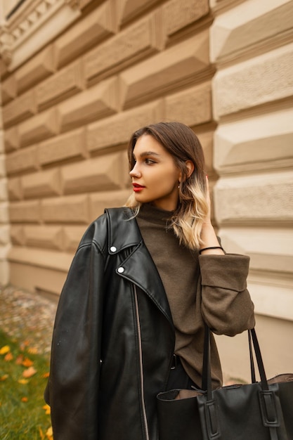 Moda joven hermosa chica en ropa casual de moda con una chaqueta de cuero y un suéter verde camina en un día de otoño cerca de un edificio antiguo