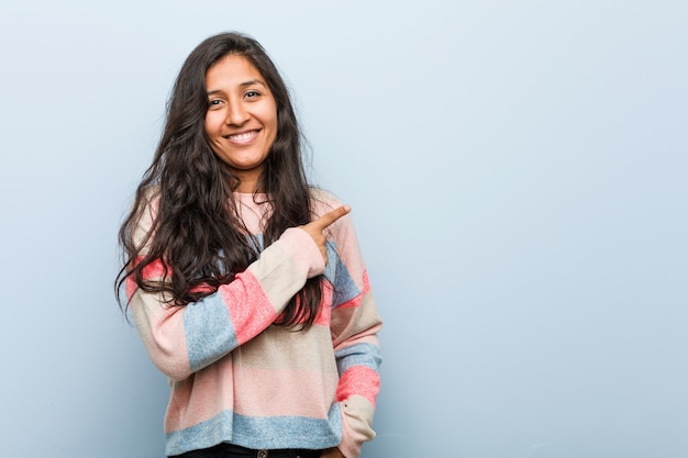 Moda joven mujer india sonriendo y apuntando a un lado, mostrando algo en el espacio en blanco.