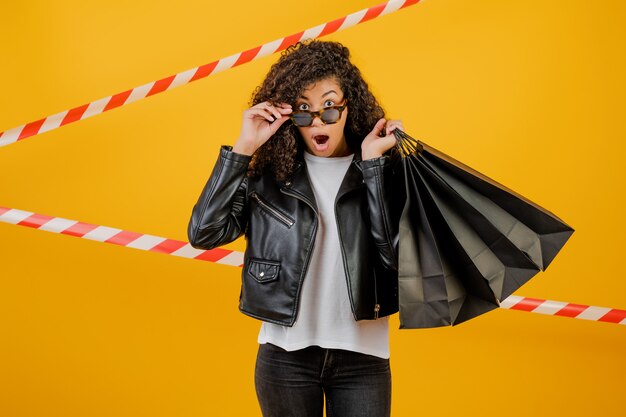 Moda joven mujer negra con chaqueta con bolsas de papel aislado sobre amarillo con cinta de señal