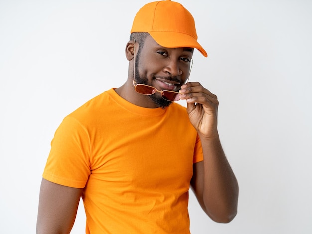 Moda masculina. Look de verão. Estilo de vida divertido. Confiante e elegante modelo de homem africano legal elegante no boné laranja combinando de t-shirt segurando óculos de sol sorrindo olhando para a câmera isolada no fundo claro.