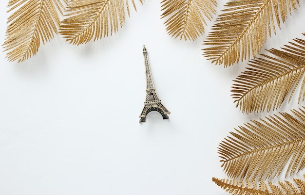 Moda minimalista ainda, vida. Estatueta da Torre Eiffel entre folhas de palmeira douradas decorativas em um fundo branco. Vista do topo