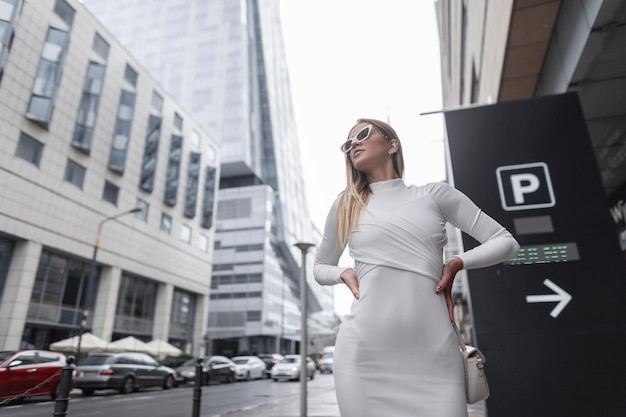 Moda modelo de mulher bonita com elegantes óculos de sol brancos em trajes da moda caminha na cidade em meio urbano