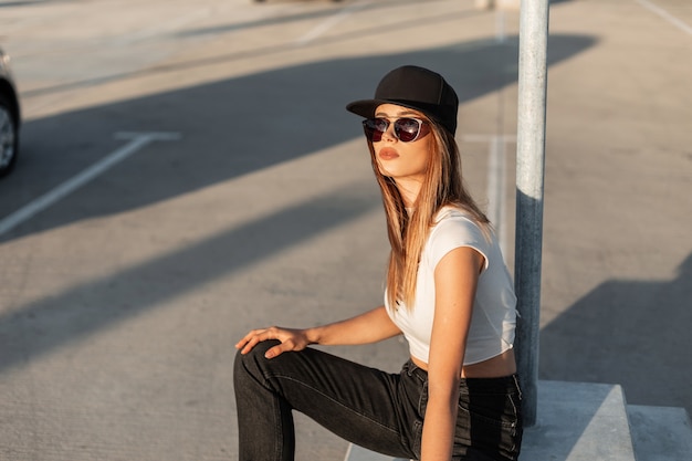 Moda modelo jovem mulher em óculos de sol pretos no boné preto legal em roupas casuais da moda de verão descansar no estacionamento e aproveitar o dia de sol. Aparência moderna americana.