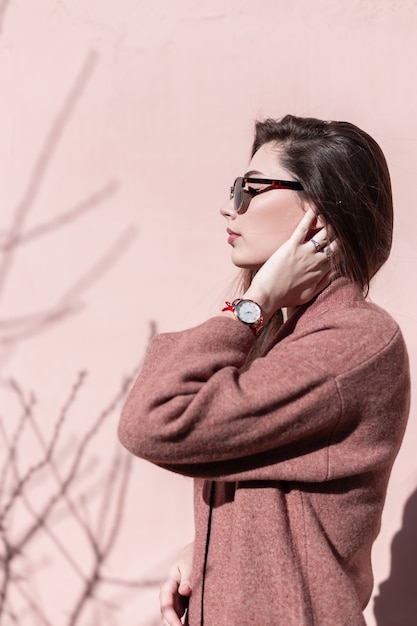 Moda modelo mulher jovem e bonita endireita o cabelo chique na rua. Elegante retrato fresco adorável garota sexy na moda óculos de sol pretos com casaco elegante vintage perto da parede rosa vintage em dia ensolarado.