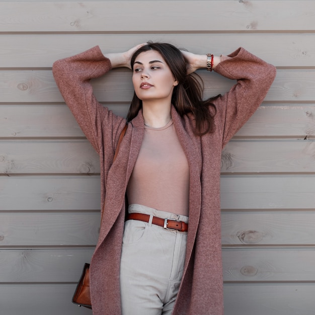 Moda modelo sexy joven elegante en ropa de moda endereza el pelo de pie junto a la pared vintage en la ciudad. Chica atractiva en abrigo en pantalones en poses de camisa en la calle cerca del edificio de madera.