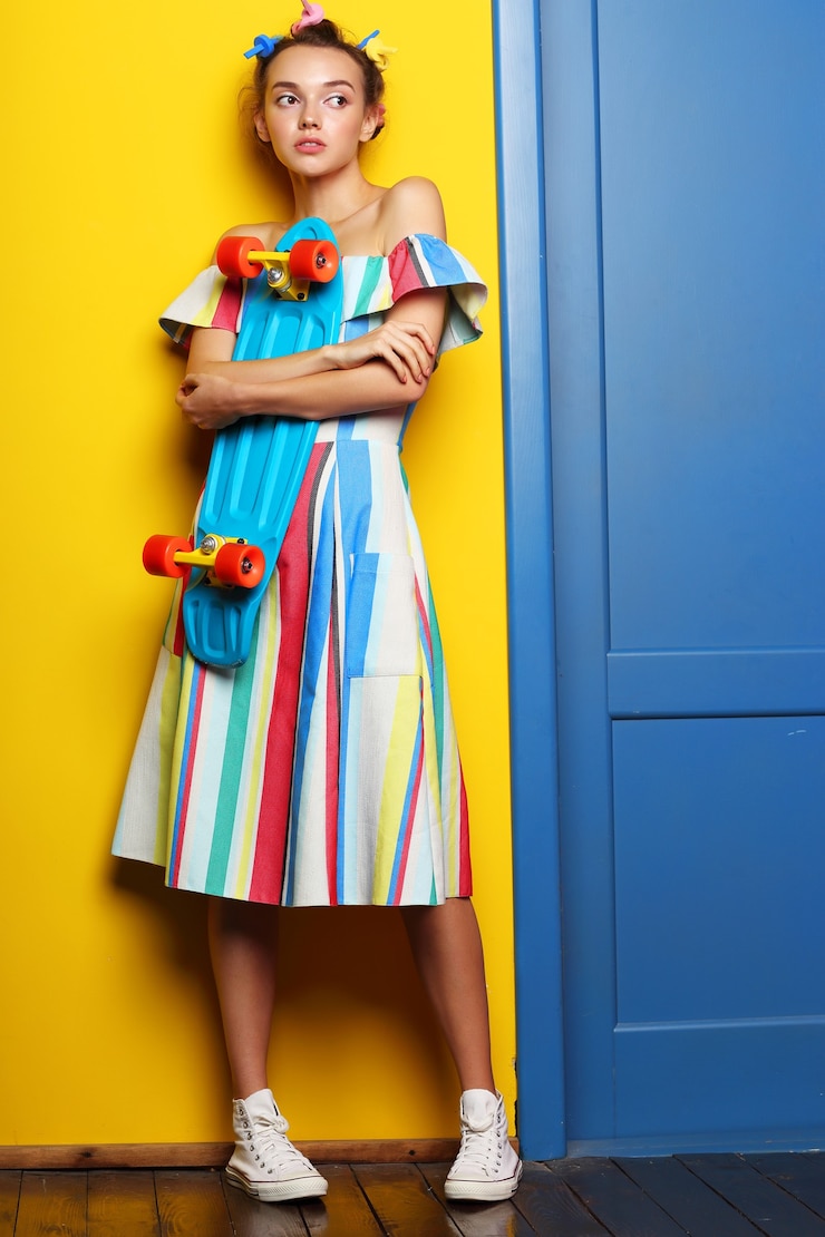 Foto moda mujer fresca posando con una patineta, vestido multicolor y zapatillas de deporte en amarillo.