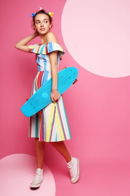 Moda mujer fresca posando con una patineta, vestido multicolor y zapatillas de deporte en rosa.