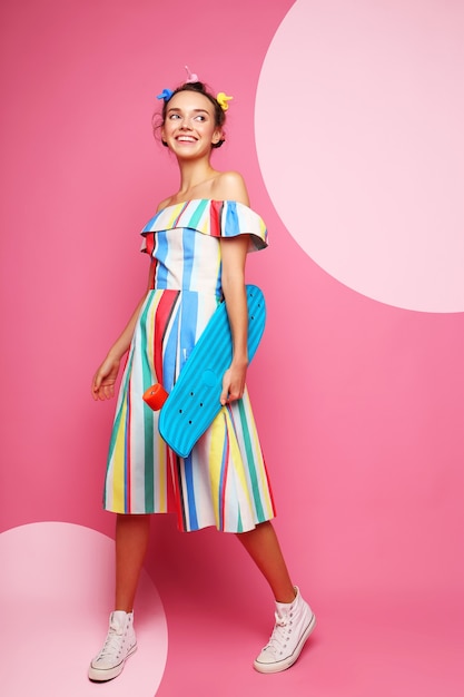 Moda mujer fresca posando con una patineta, vestido multicolor y zapatillas de deporte en rosa.