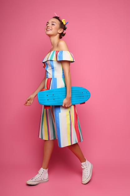 Moda mujer fresca posando con una patineta, vestido multicolor y zapatillas de deporte en rosa.