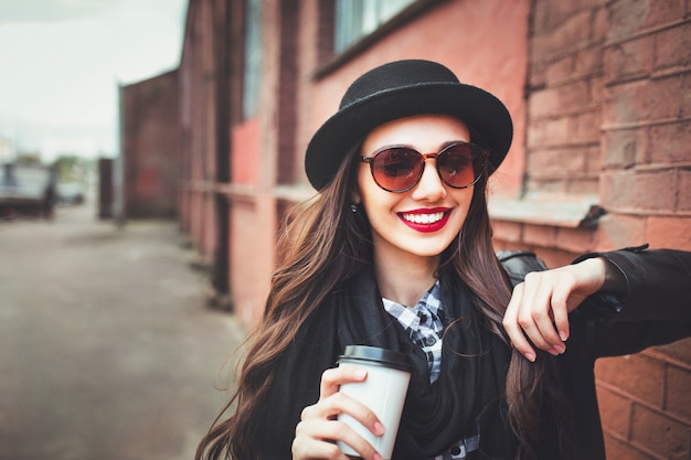 Moda mujer en gafas de sol y sombrero con bebida al aire libre. Mujer joven en gafas de sol en la calle y sosteniendo la taza de café