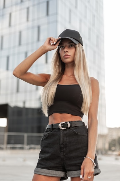 Moda mujer hermosa en una elegante camiseta negra y pantalones cortos de jeans negros con una gorra camina en la ciudad