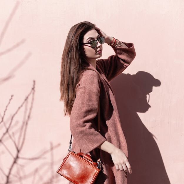 Moda mujer joven atractiva con hermoso cabello largo con labios sexy en gafas de sol de moda con bolso posando de pie en el sol junto a la pared rosa vintage en la ciudad. Elegante modelo de niña dulce en ropa elegante