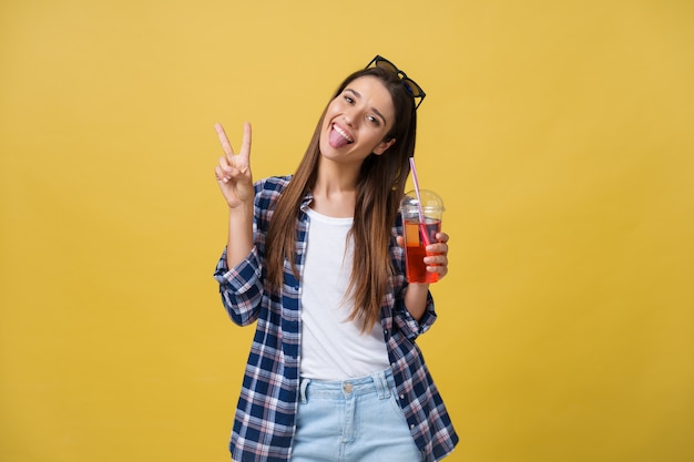 Moda mujer joven y bonita con taza de jugo de fruta fresca en camisa azul divirtiéndose sobre fondo amarillo colorido