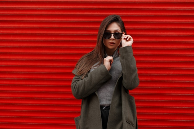 Moda mujer joven hermosa con un abrigo de moda y un suéter gris endereza elegantes gafas de sol cerca de la pared de metal rojo