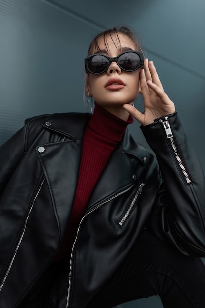 Moda mujer joven hermosa con gafas de sol frescas en elegante chaqueta de cuero negro con suéter burdeos posando en la ciudad