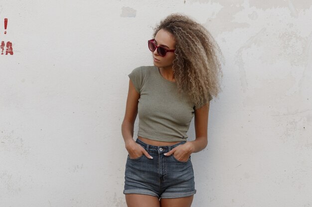 Moda mujer joven hermosa modelo sexy con pelo rizado en una camiseta y pantalones cortos de mezclilla con estilo se encuentra cerca de una pared blanca en la calle