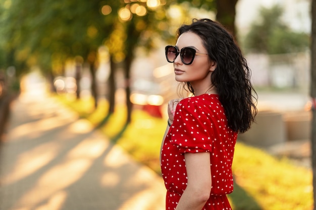 Moda mujer joven inconformista con gafas de sol en vestido rojo de moda caminando por la calle al atardecer