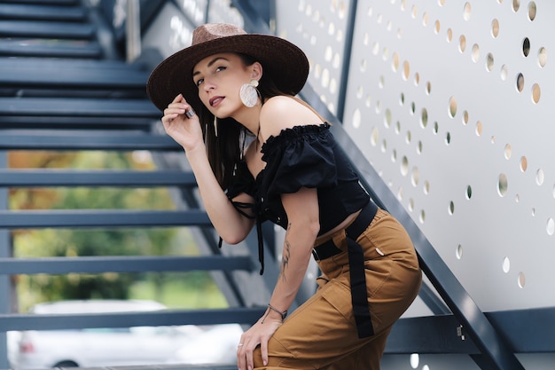 Moda mujer morena con cabello largo, con elegante sombrero de mimbre grande, posando en las escaleras.