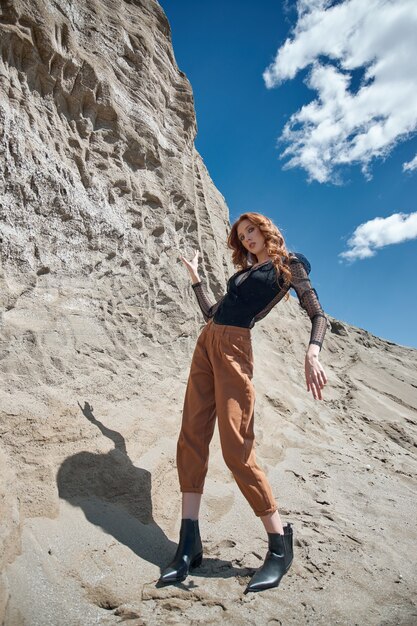 Moda mujer pelirroja en jeans naranja posando en la naturaleza cerca de rocas arenosas