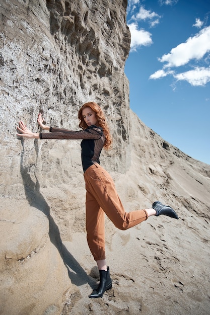 Moda mujer pelirroja en jeans naranja posando en la naturaleza cerca de rocas arenosas