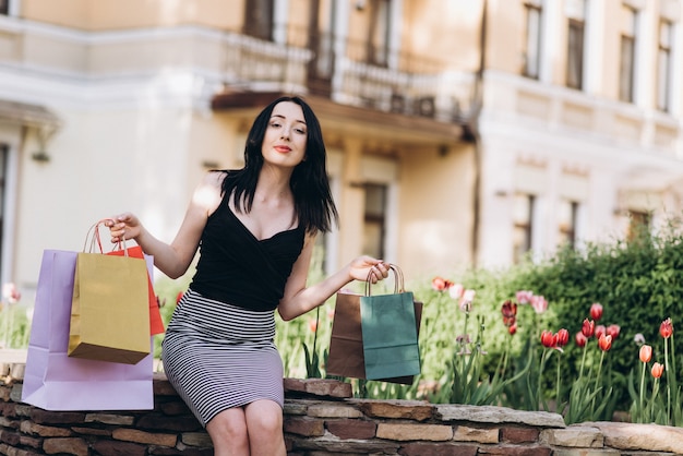 Moda mujer vestida con bolsas de compra de color sentado cerca de flores en las calles, concepto de compras