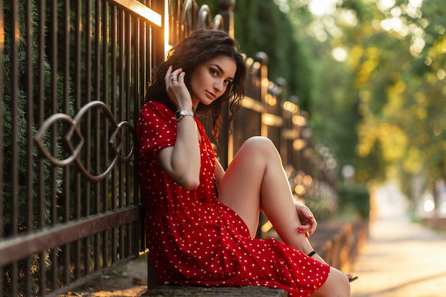 Moda mulher bonita com cabelo encaracolado em um elegante vestido vermelho sentada perto de uma cerca na cidade