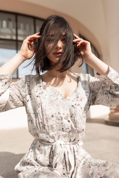 Moda mulher de beleza muito jovem com penteado curto no vestido de flores elegantes da primavera senta-se na rua em um dia ensolarado e ventoso