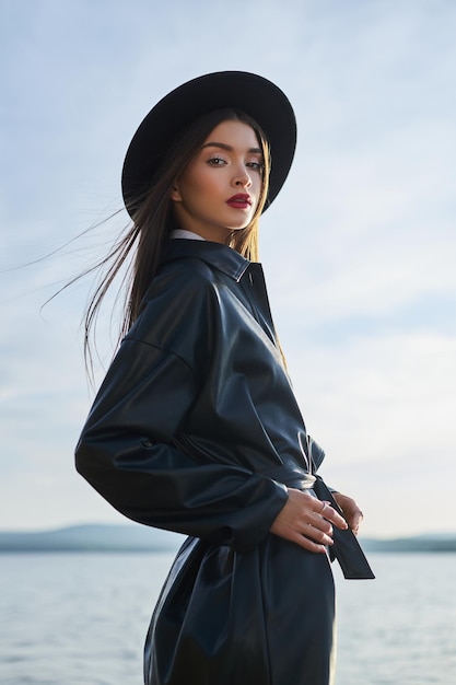 Moda mulher de chapéu preto redondo e capa de chuva de couro perto da lagoa do lago. menina do retrato ao pôr do sol. bela maquiagem e batom vermelho brilhante nos lábios