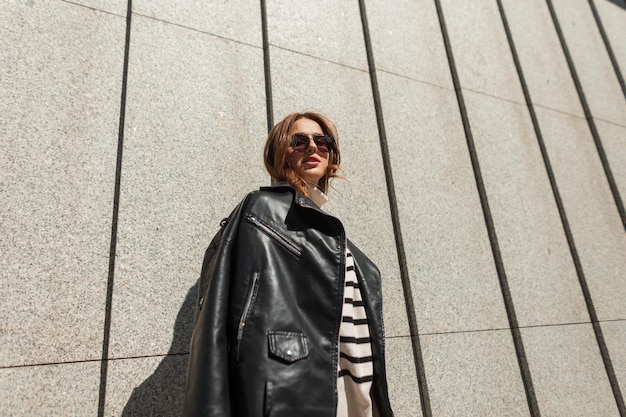 Moda mulher jovem e bonita com óculos de sol da moda em roupas elegantes urbanas com uma jaqueta de couro preta com um suéter listrado perto da parede na rua em um dia ensolarado
