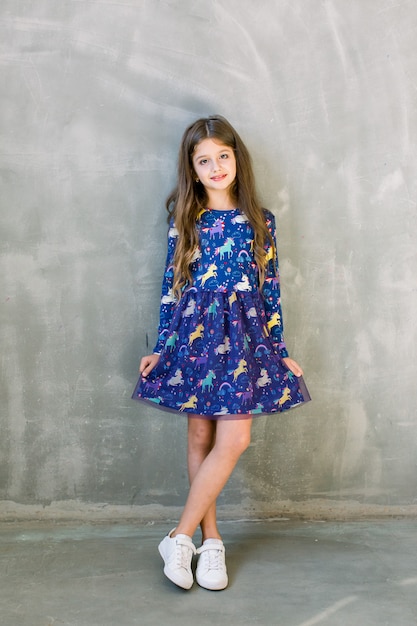 Moda niña pequeña en vestido azul en una pared gris