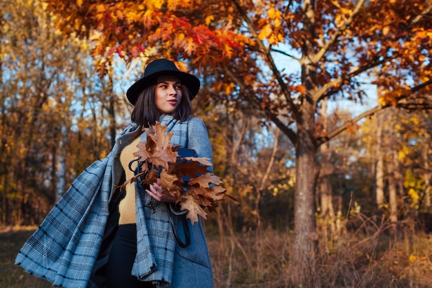 Moda outono. Mulher jovem caminhando na floresta, vestindo roupas elegantes e segurando a bolsa com folhas. Roupas e acessórios