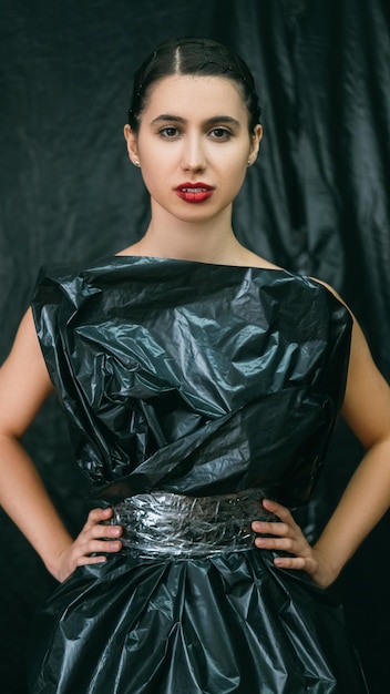 Moda plástica Reciclaje de residuos Colección ecológica Protección del medio ambiente Mujer segura con maquillaje nocturno posando en vestido negro de bolsa de basura aislado sobre fondo oscuro de polietileno arrugado