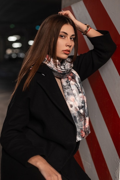 Moda retrato elegante jovem mulher com casaco preto elegante em xale de seda na moda perto do pilar vermelho e branco moderno na rua perto da estrada. Modelo de mulher bonita moderna com roupas da moda ao ar livre.