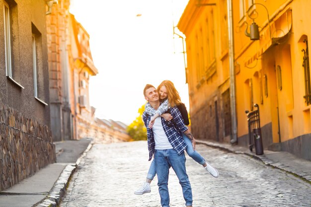 Moda verano imagen joven pareja con estilo en la vieja ciudad romántica. Hombre que sostiene a la mujer joven a cuestas en el fondo de los edificios al atardecer