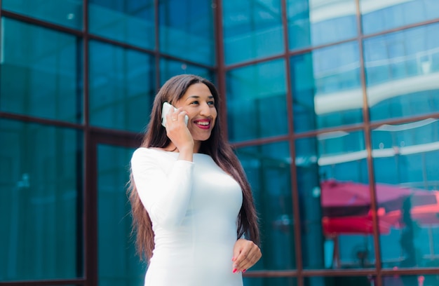 Mode-Afro-Frau in einem luxuriösen weißen Kleid, das am Telefon gegen Gebäude mit blauen Fenstern im Freien spricht.