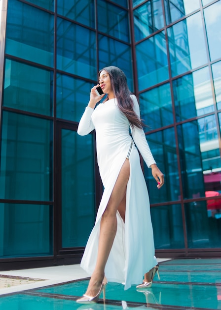 Mode-Afro-Frau in einem luxuriösen weißen Kleid, das am Telefon spricht, während gegen Gebäude mit blauen Fenstern im Freien geht.