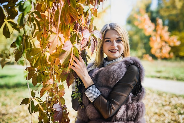 Mode Frau. Lächelndes Mädchen in Pelzmantel Posin im Herbstpark mit Bäumen und Efeu