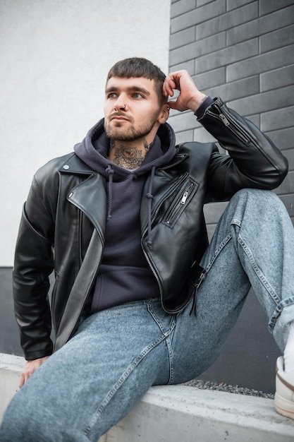 Mode Hipster gutaussehender Mann mit Tätowierungen am Hals in Freizeitkleidung mit Lederjacke Hoodie und Jeans sitzt und posiert in der Nähe einer weißen Backsteinmauer auf der Straße