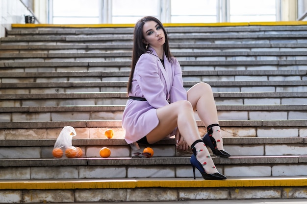 Mode hübsches Frauenmodell mit Fruchtorangen sitzt auf den Stufen in einer lila Jacke, schwarze Schuhe.