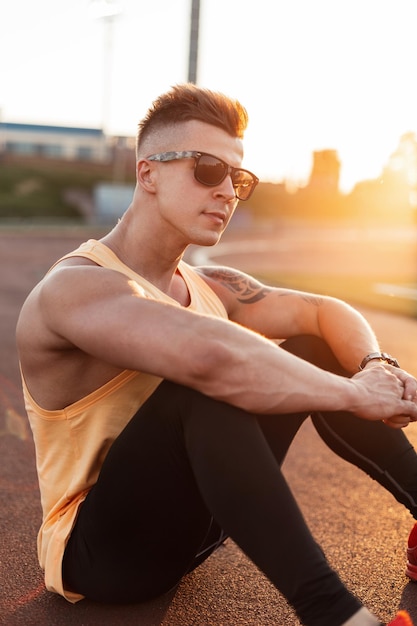 Mode junger Hipster-Fitness-Mann mit Sonnenbrille in Sportbekleidung sitzt bei Sonnenuntergang im Stadion