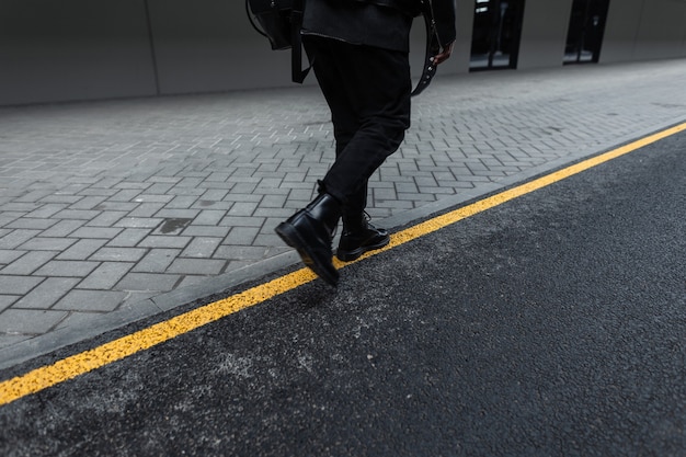 Mode junger Mann in einer stilvollen Jacke in Vintage-Jeans in modischen Lederstiefeln mit einem trendigen schwarzen Rucksack geht draußen auf der Straße spazieren. Cooler Typ in schwarzer Jugendkleidung. Rückansicht. Gelbe Linie