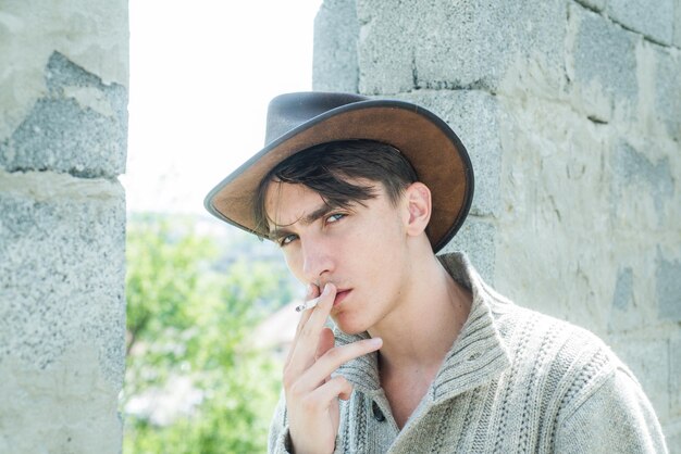 Mode junger Mann. Schöner Kerl mit selbstbewusstem Gesicht. Nahaufnahme junger Mann, der eine Zigarette raucht. Mann mit Zigarre. Gut aussehender Mann trägt Cowboyhut.