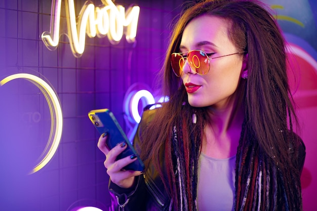 Mode-Mädchen, das ihr Smartphone beim Stehen in der Nähe von Leuchtreklamen im Nachtclub verwendet