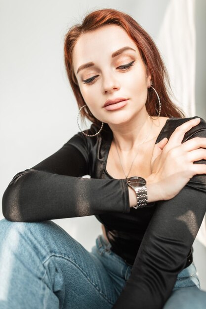 Mode-Model der jungen Frau mit süßem Gesicht und sauberer Haut in stylischem schwarzem T-Shirt und blauer Jeans sitzt drinnen bei Sonnenlicht