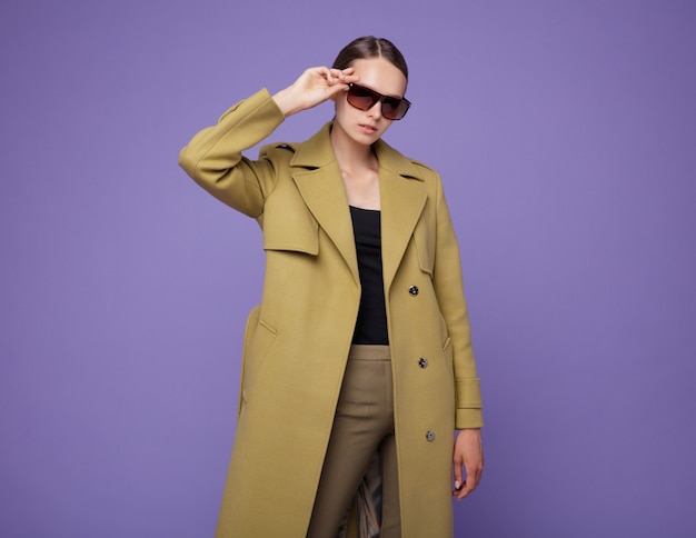 Mode-Modell im gelben Senf-Umhang auf lila Hintergrund Studioaufnahme Porträt Stilvolle Sonnenbrille