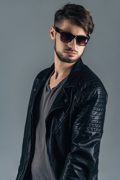 Mode-Modell. Selbstbewusster junger Mann mit Sonnenbrille, der wegschaut, während er vor grauem Hintergrund steht